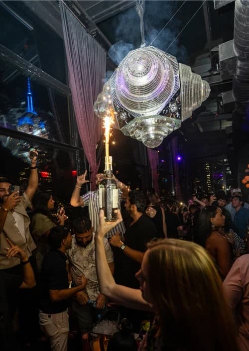 Crowded rooftop bar at Suave Saturdays in Manhattan, with a bottle of champagne being served with a sparkler and people enjoying the music and atmosphere.