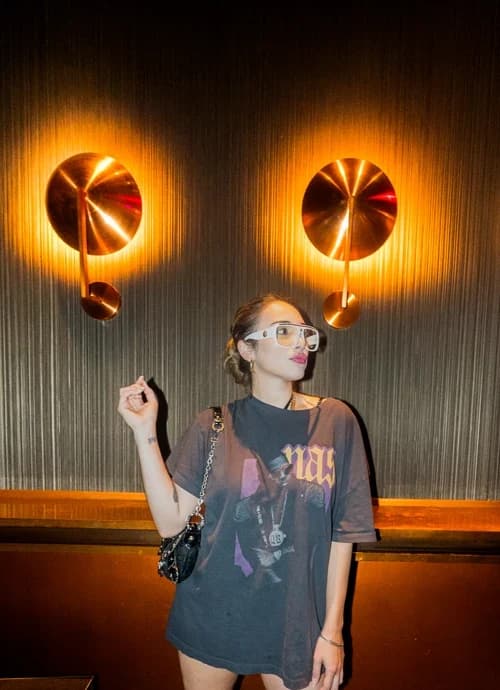 Woman posing stylishly in an oversized t-shirt and white sunglasses against a wall with modern light fixtures at Suave Saturdays.